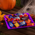 Paarse kom met diverse Halloween-chocoladerepen, een pompoen en spinnenwebben op een houten tafel.